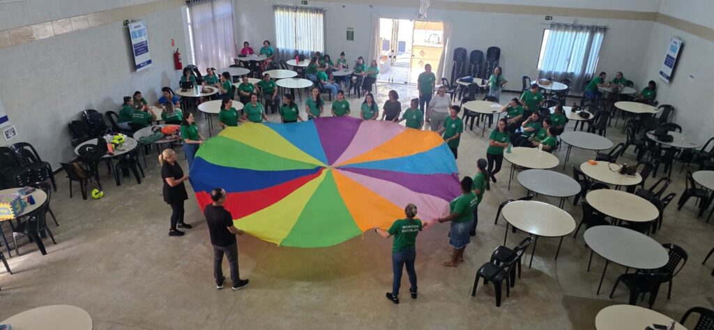 Grupo de profissionais de apoio escolar participando de dinâmica com paraquedas colorido em oficina prática